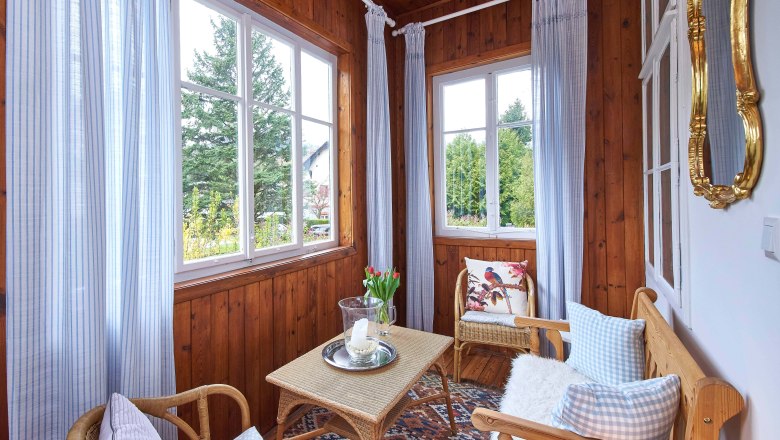 Ein gemütlicher Wintergarten mit Holzwänden, Korbmöbeln, blauen Vorhängen und einem Tisch mit Kerze und Blumen.