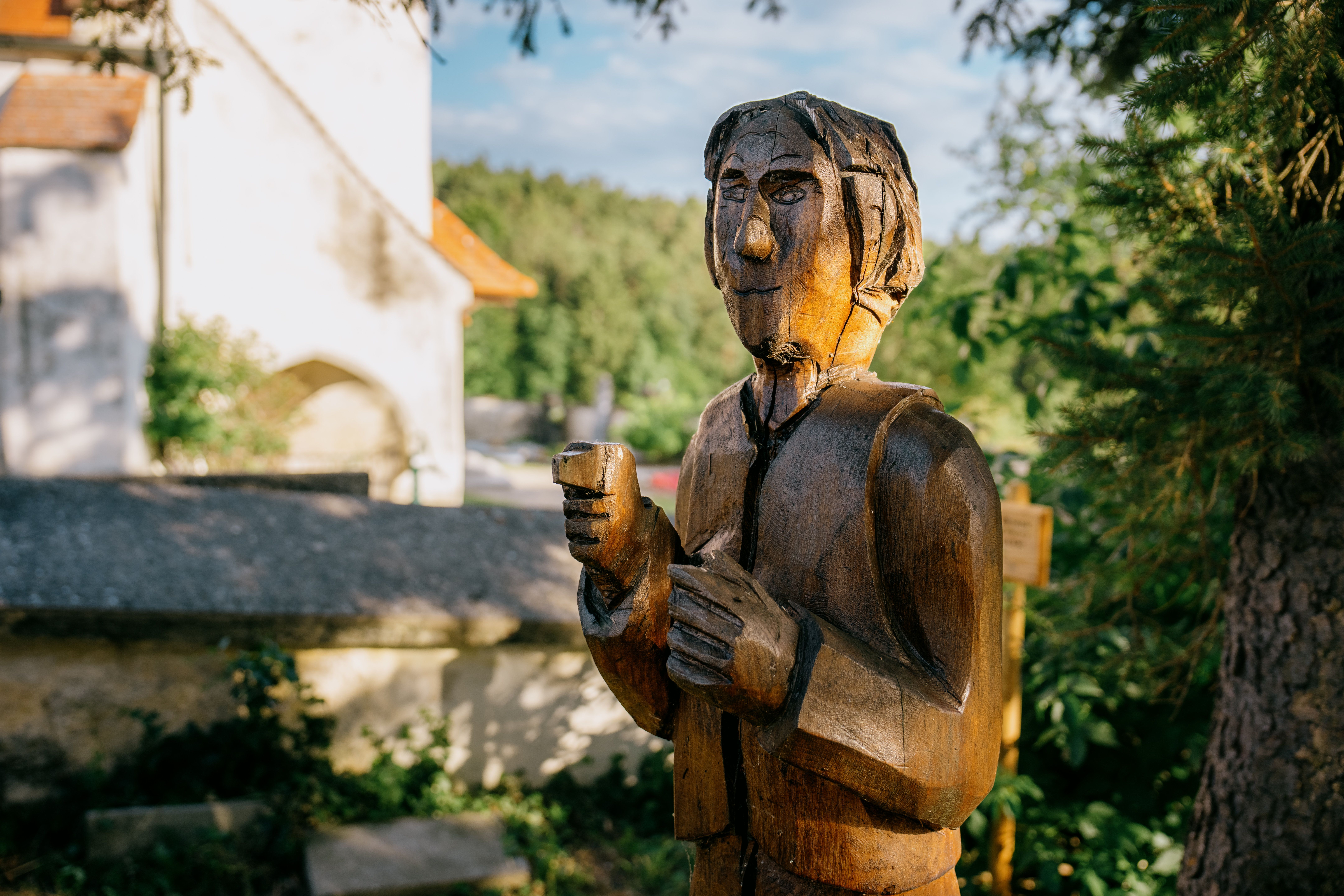 A wooden figure of a legendary character on the Lanzenkirchen Legend Trail, in front of the church walls in Ofenbach and surrounded by green nature.