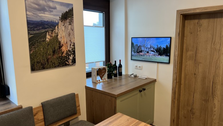 Kitchen with wooden furniture, a landscape painting on the wall and a TV showing a picture of a house.