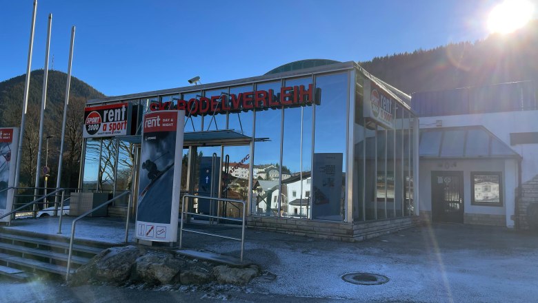 Außenansicht eines Skiverleihs mit Glasfassade und Werbeschildern, umgeben von Bergen und strahlendem Sonnenschein.