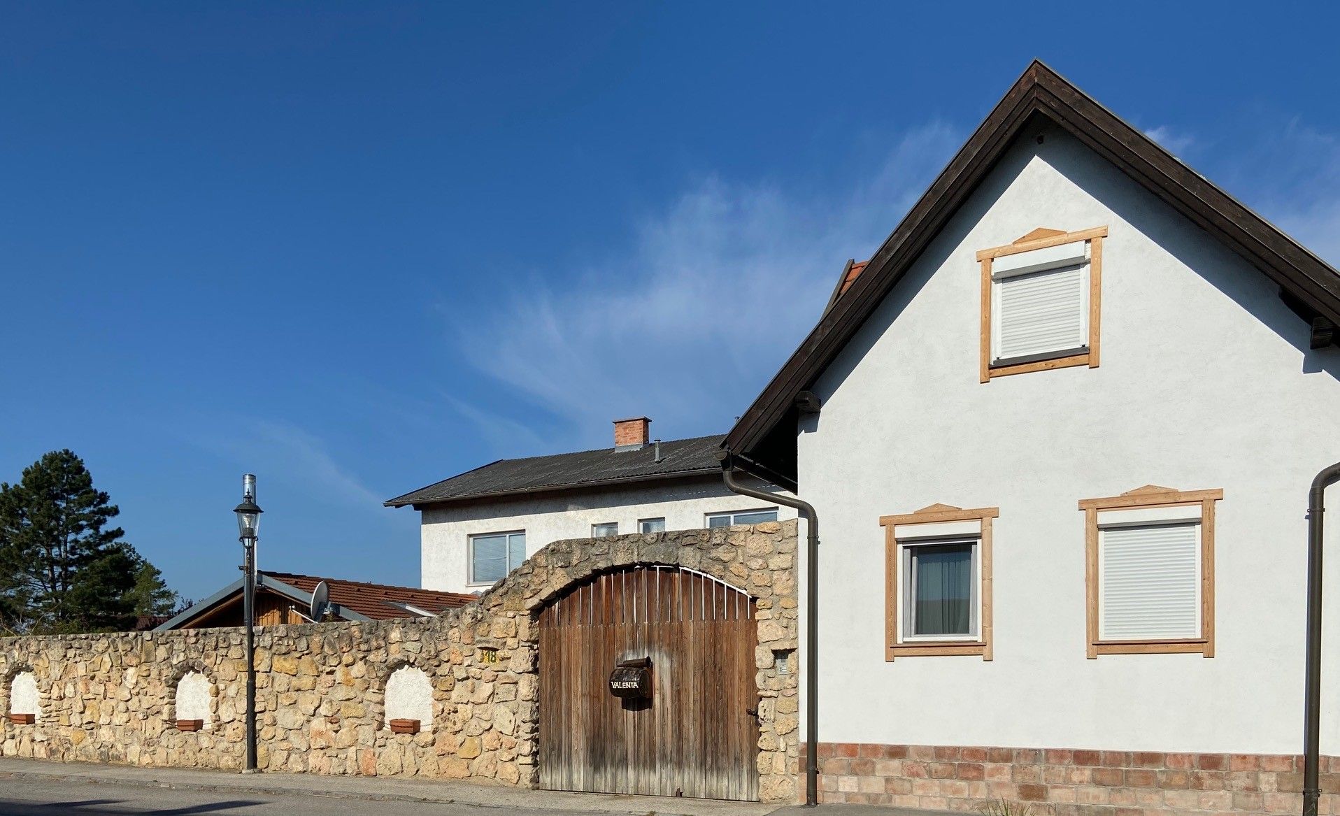 Ein weißes Haus mit braunen Fensterrahmen und einer Steinmauer mit Holztor unter blauem Himmel.