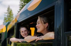 Zwei fröhliche Frauen schauen aus dem Fenster des Salamander-Zuges der Schneebergbahn, während die Landschaft aus Bäumen im Hintergrund vorbeizieht, was Freude und Neugier auf das bevorstehende Reiseerlebnis widerspiegelt.
