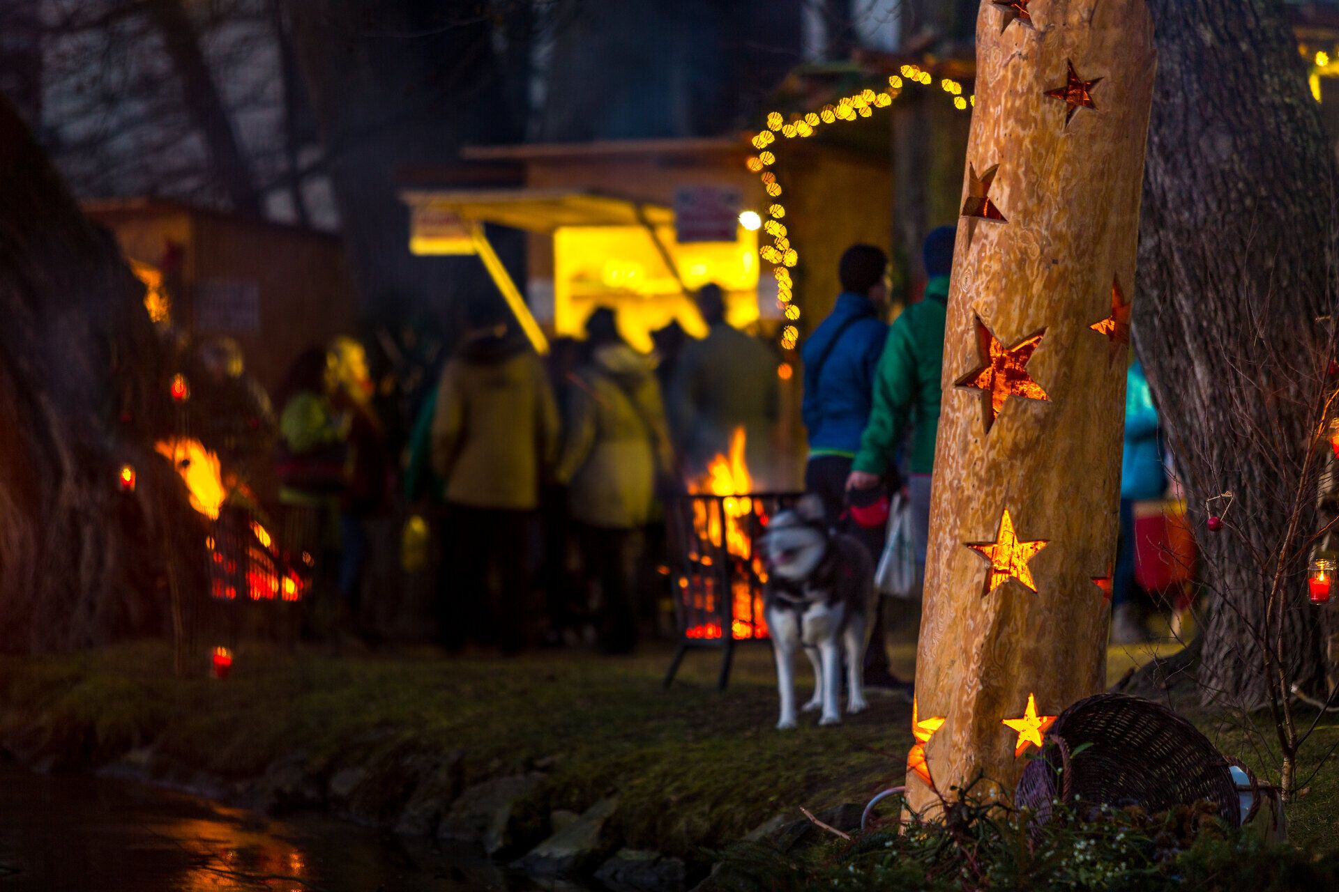 Personen auf einem stimmungsvoll beleuchteten Adventmarkt mit Feuerschalen und einer Holzskulptur mit leuchtenden Sternen.