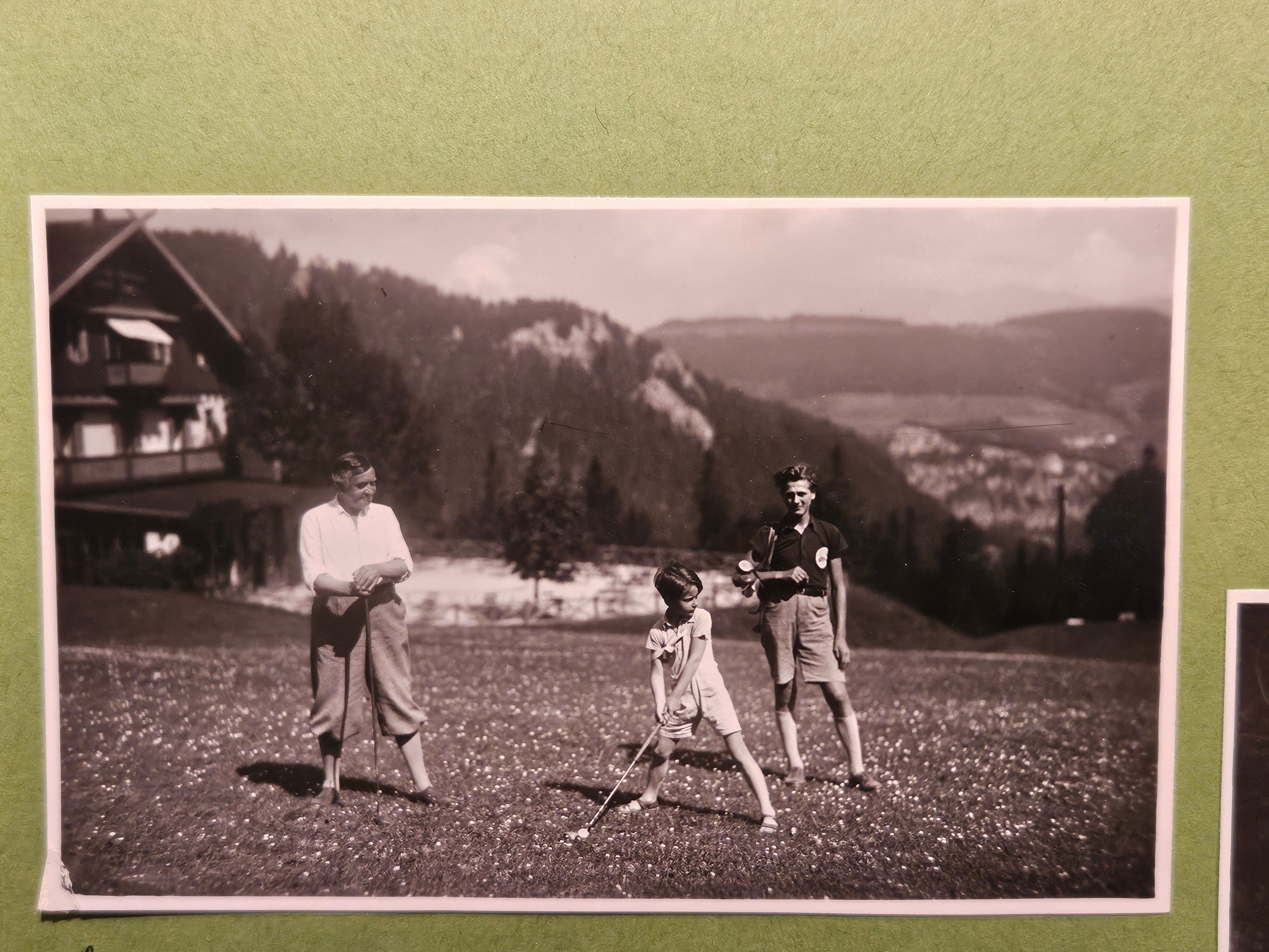 Ein Erwachsener und zwei Kinder spielen auf einem Golfplatz in alpiner Landschaft und einem Gebäude im Hintergrund.
