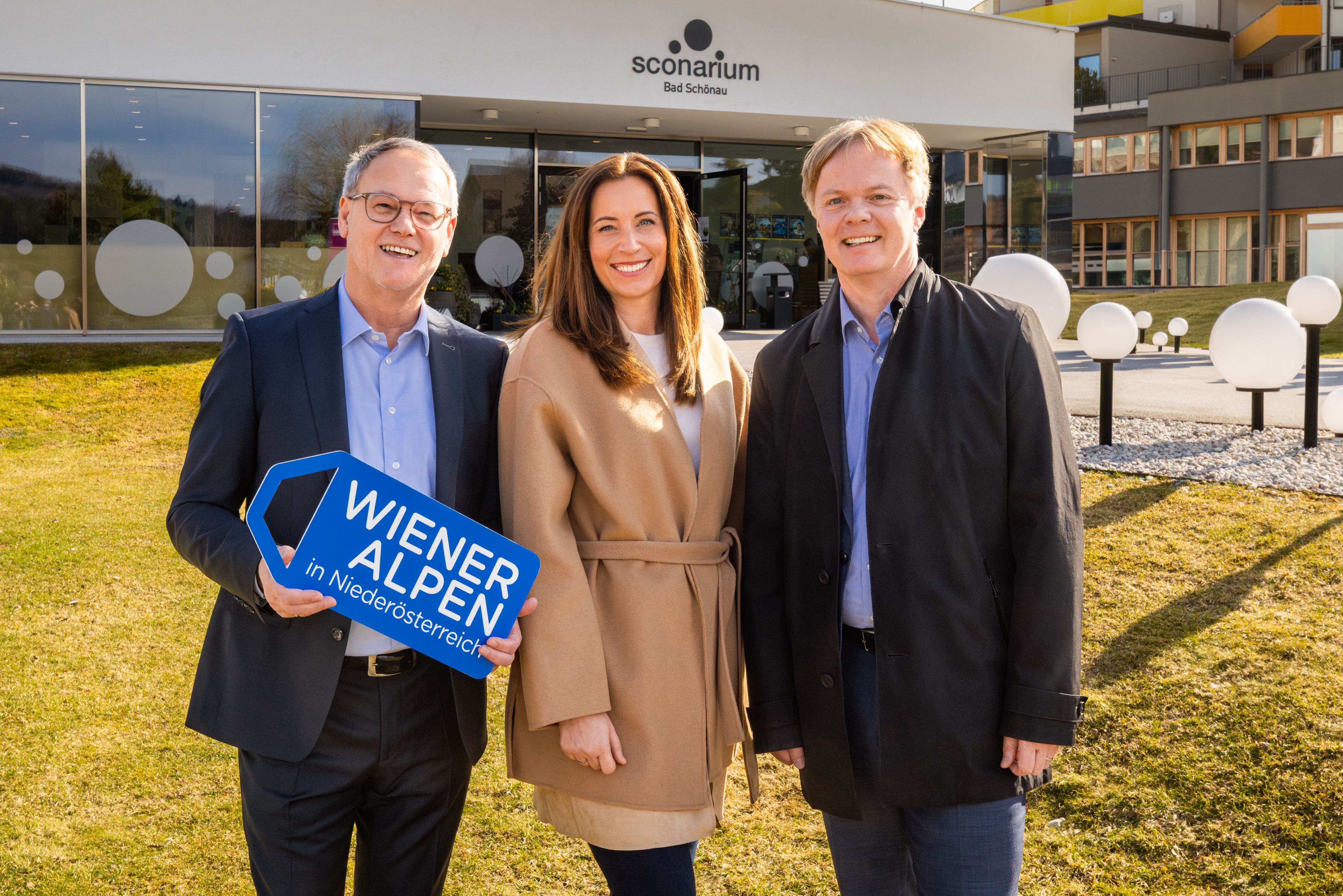 Drei Personen stehen vor dem Kulturhaus, einem modernen Gebäude mit Glasfront und der Aufschrift "Sconarium Bad Schönau". Der Bürgermeister hält ein das Logoschild "Wiener Alpen" in den Händen. 
