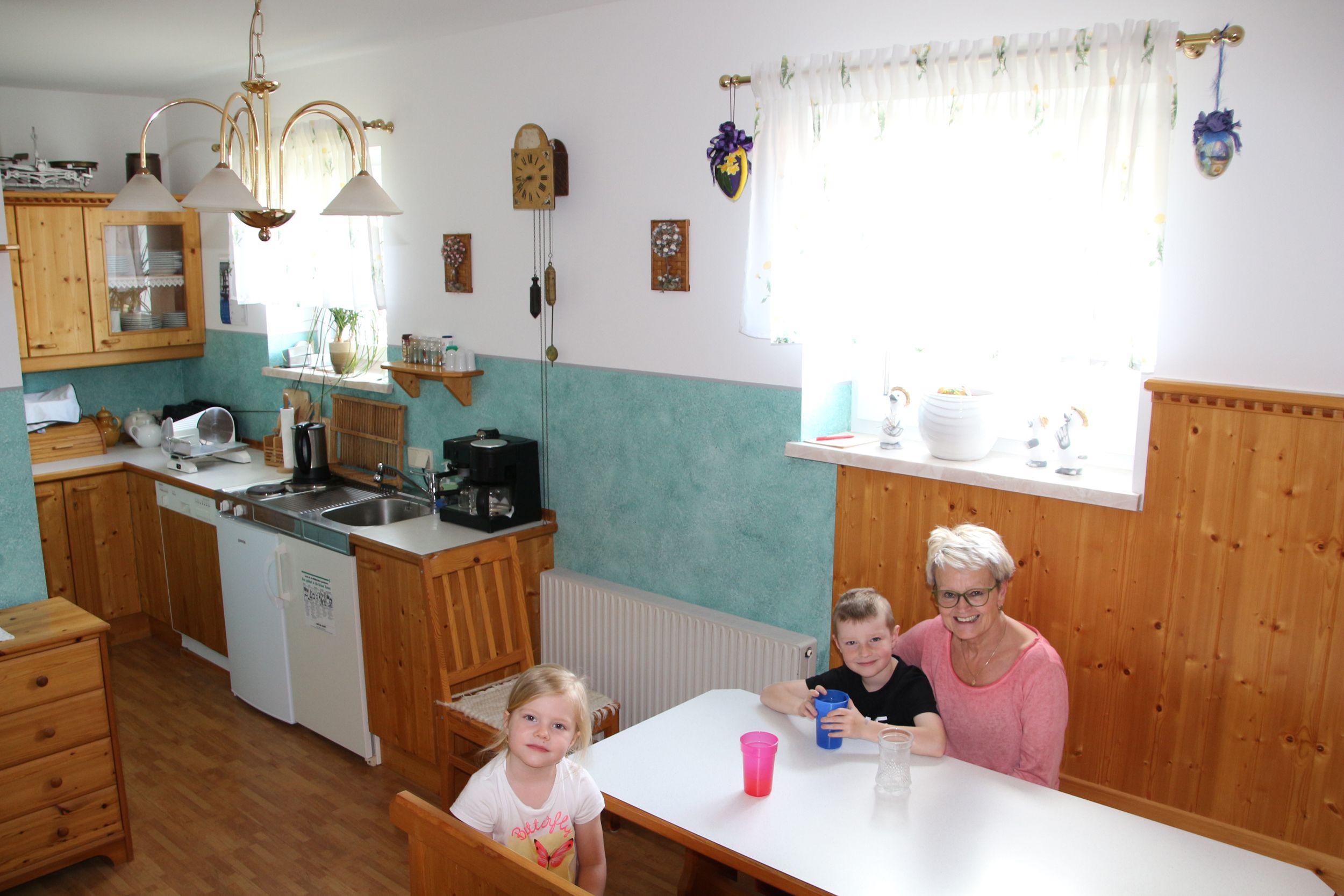 Ein Frühstücksraum mit Holzmöbeln, einer Küche im Hintergrund und drei Personen an einem Tisch.