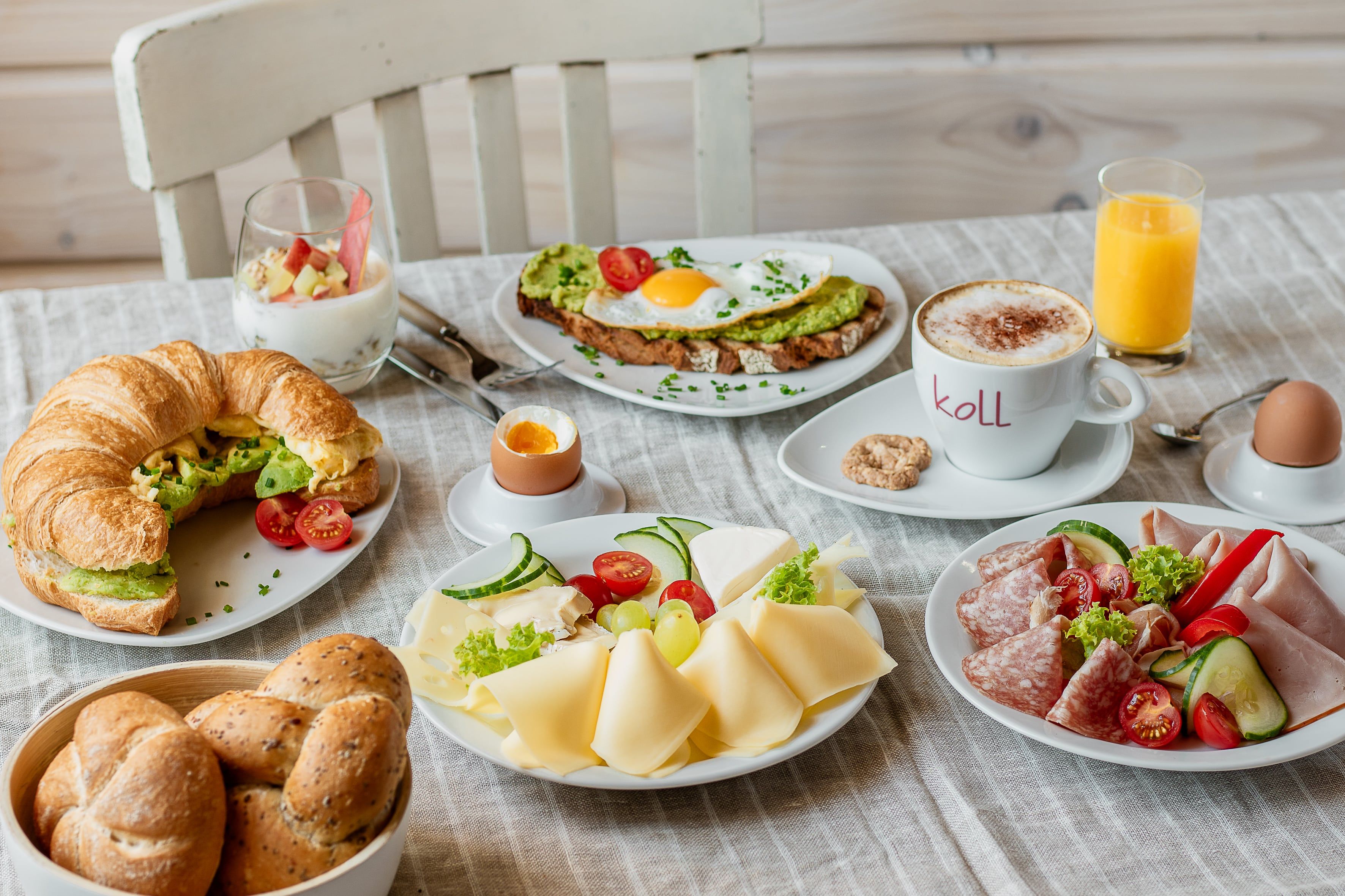 Ein reichhaltiges Frühstück mit Croissant, Brot, Käse, Aufschnitt, Ei, Joghurt, Kaffee und Orangensaft auf einem Tisch.