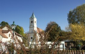Pfarrkirche Walpersbach mit Turm und umliegenden Gebäuden, umgeben von Bäumen und Sträuchern unter blauem Himmel.