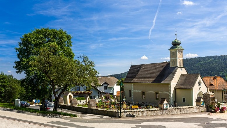 Small church with tower in a village, surrounded by trees and houses, under a blue sky.