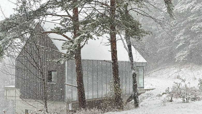 Ein modernes Haus im Wald, umgeben von Schnee und Bäumen, während es stark schneit.