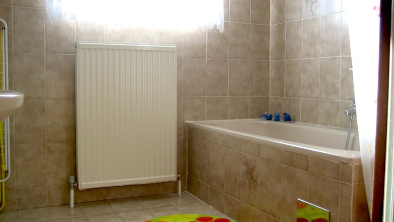 Ein Badezimmer mit beiger Fliesenwand, Badewanne, Heizkörper und bunten Teppichen.