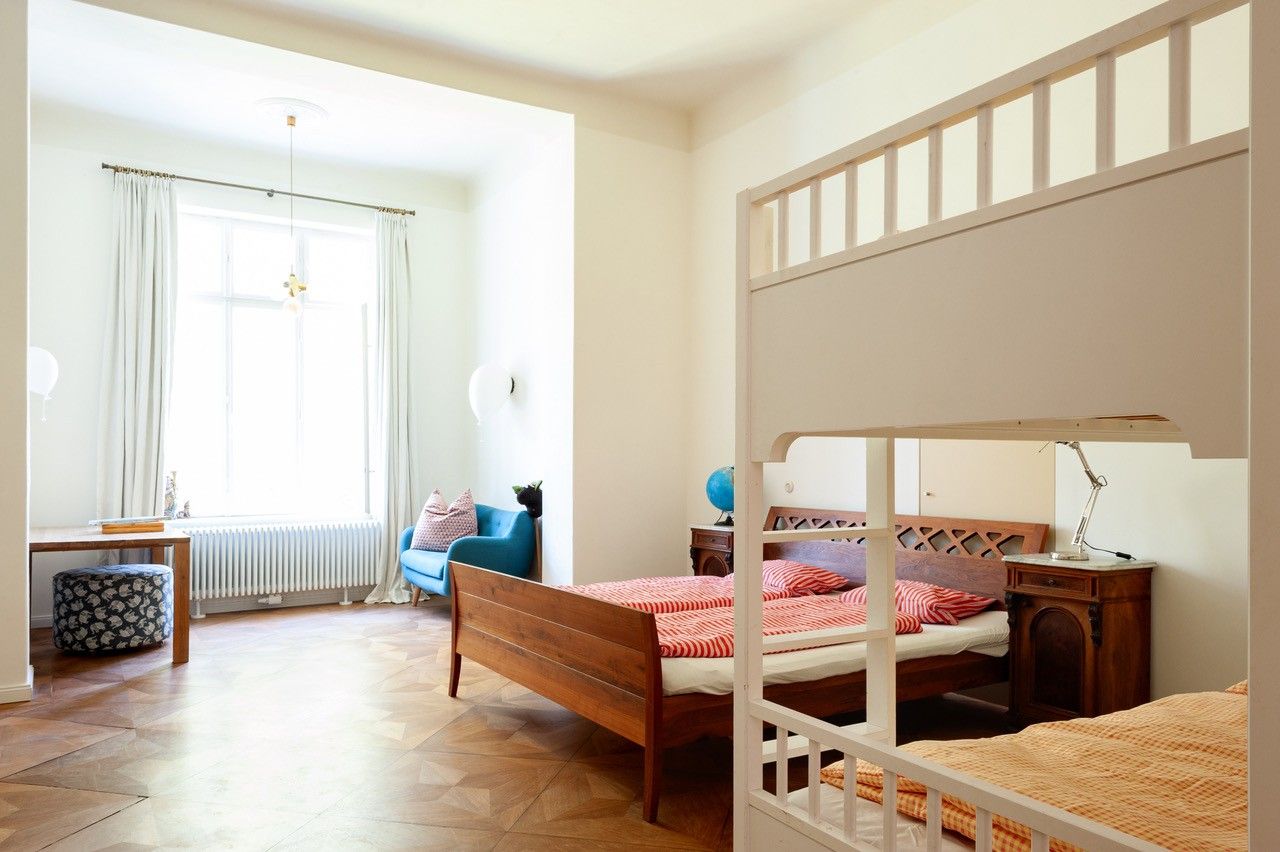 Ein helles Schlafzimmer mit einem Etagenbett, einem Einzelbett, einem blauen Sessel und einem Schreibtisch vor einem großen Fenster.