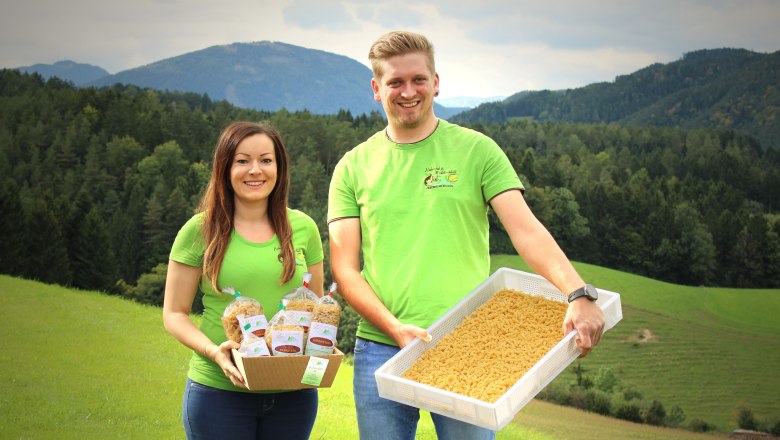 Zwei Personen in grünen T-Shirts präsentieren Nudeln vor einer Berglandschaft.