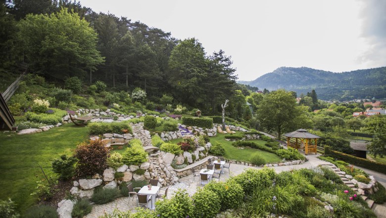 A terraced garden with stones, plants and seating areas, surrounded by trees and mountains in the background.
