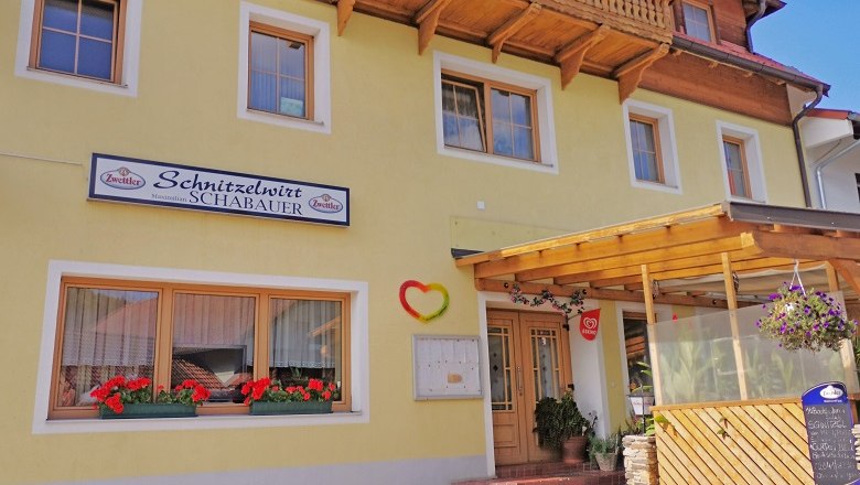 Gelbes Gebäude mit Schild 'Schnitzelwirt Schabauer', Blumenkästen und überdachtem Eingang.