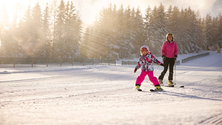 Child and adult skiing in the snow, surrounded by trees and sunshine.