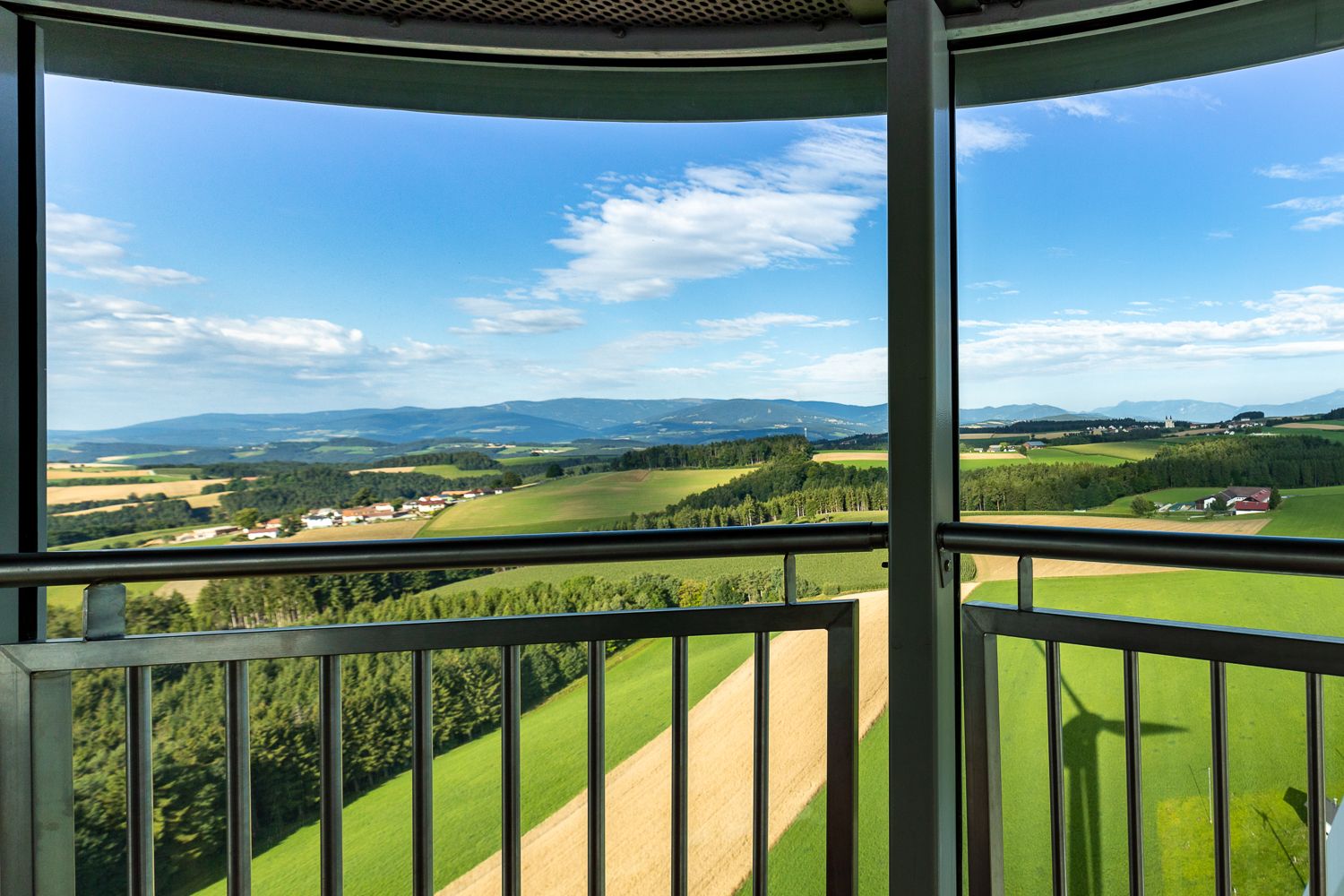 Aussicht von der Kanzel des Windrades aus auf eine grüne Landschaft mit Feldern und Hügeln unter blauem Himmel.