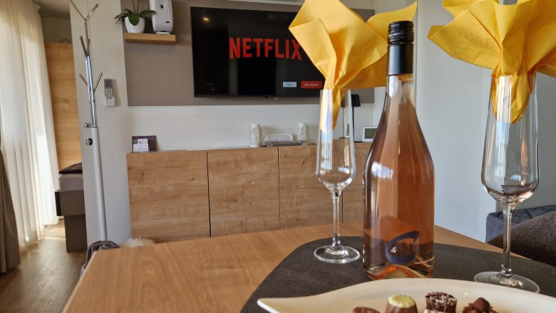 Interior view of a chalet with a bottle of wine, glasses and chocolates on a table, television in the background.
