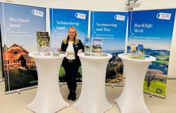 Vor vier Rückwänden einzelner Regionen der Wiener Alpen steht eine Frau hinter drei Stehtischen, auf denen verschiedene Broschüren angeboten werden. 