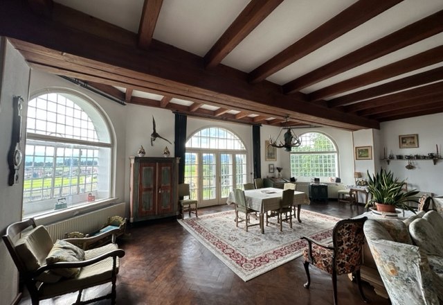 Ein elegantes Wohnzimmer mit Holzbalkendecke, großen Fenstern, antiken Möbeln und einem Teppich.