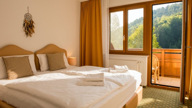 Ein gemütliches Doppelzimmer mit zwei Betten, einem Balkon mit Blick auf bewaldete Hügel und einem Traumfänger an der Wand.