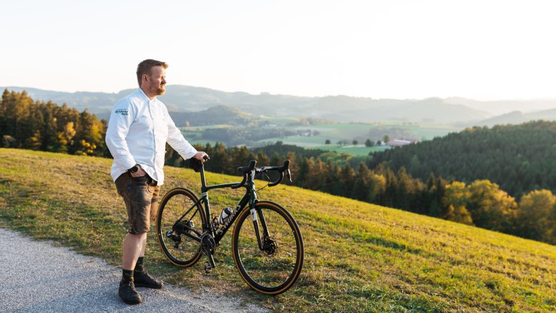 Ein Mann in weißem Hemd steht mit einem Fahrrad auf einem Hügel mit Blick auf eine grüne Landschaft.