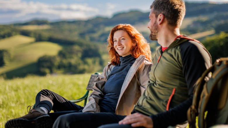 Zwei Personen sitzen auf einer Bank in einer grünen Landschaft.