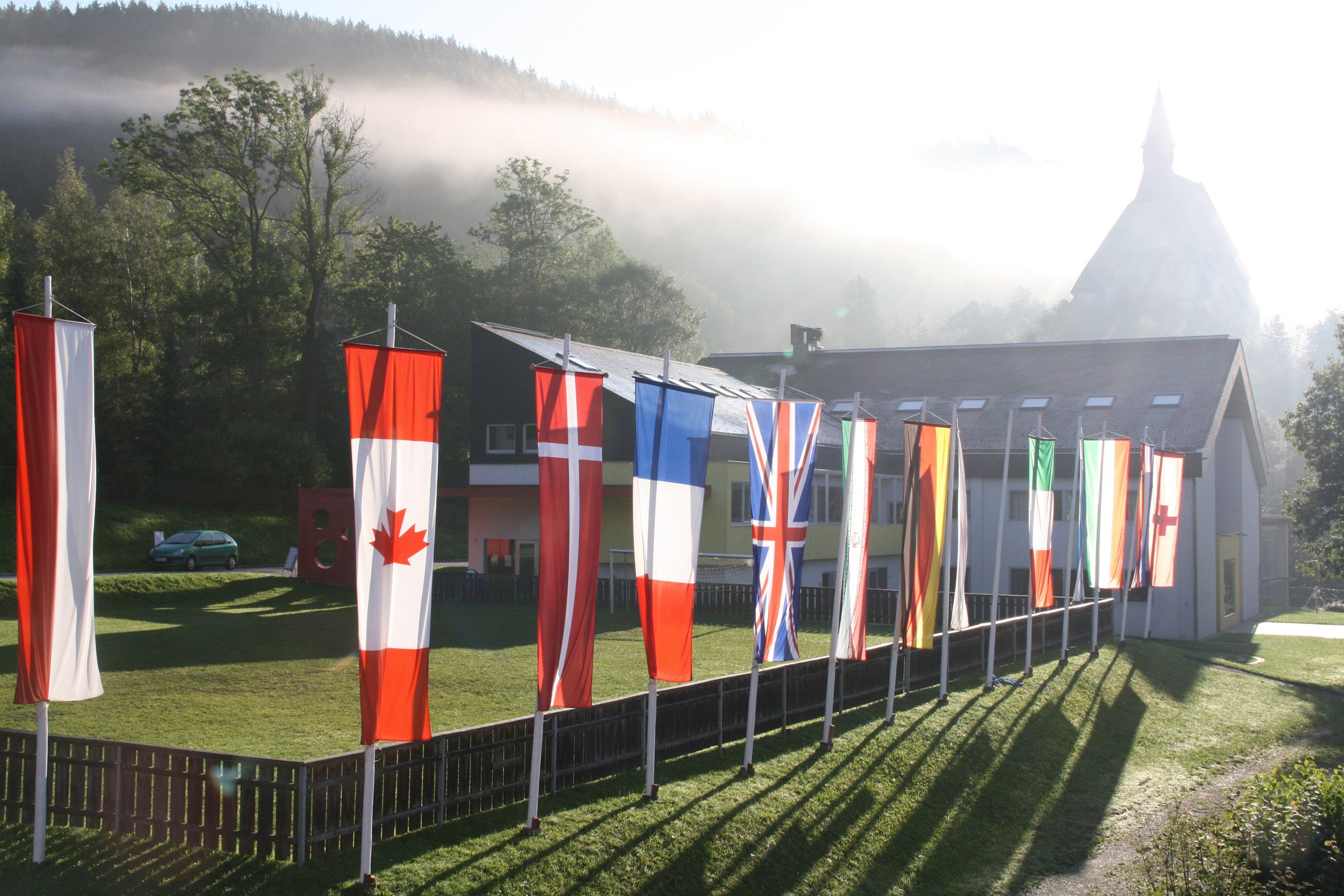Verschiedene Nationalflaggen vor einem Gebäude in einer ländlichen Umgebung mit Nebel und Bäumen.