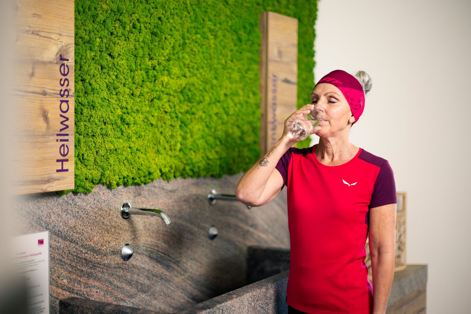 Eine sportlich gekleidete Frau nimmt einen Schluck Wasser aus einem Glas vor Wasserhähnen und der Beschriftung "Heilwasser".