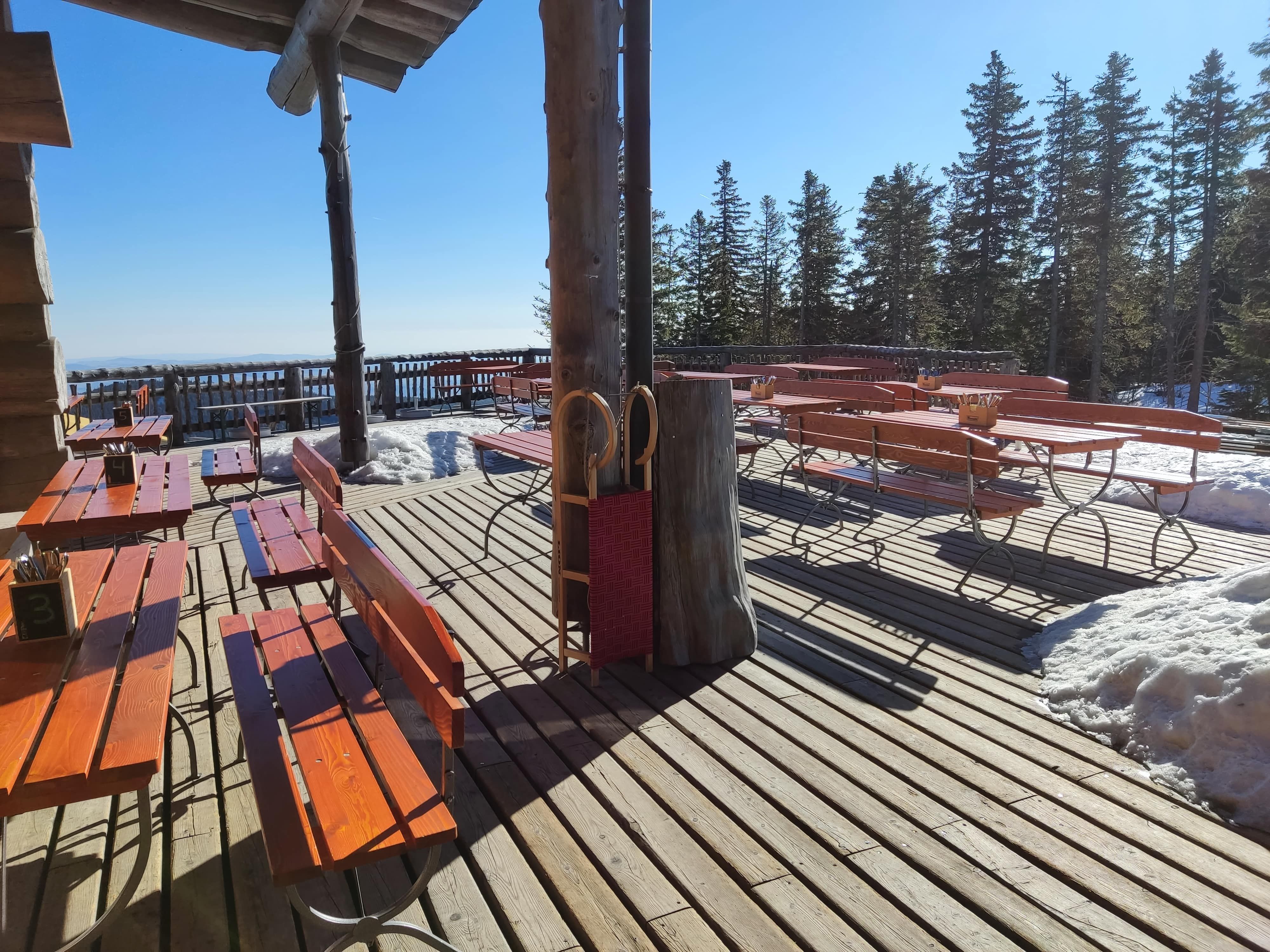 Holzterrasse mit Bänken und Tischen, umgeben von Schnee und Bäumen.