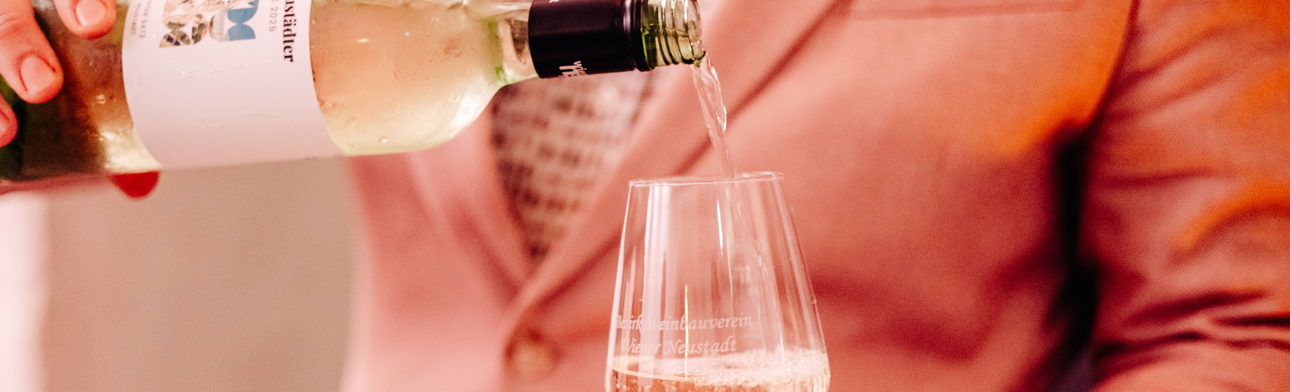 In ein Weinglas wird ein Weißwein eingeschenkt. Im Hintergrund sieht man einen Mann mit rosa Sakko. 