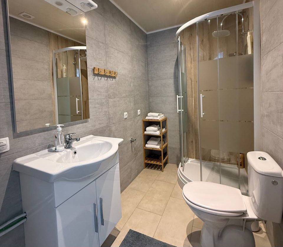 Modernes Badezimmer mit Dusche, Waschbecken und Toilette. Handtücher sind auf einem Regal gestapelt.