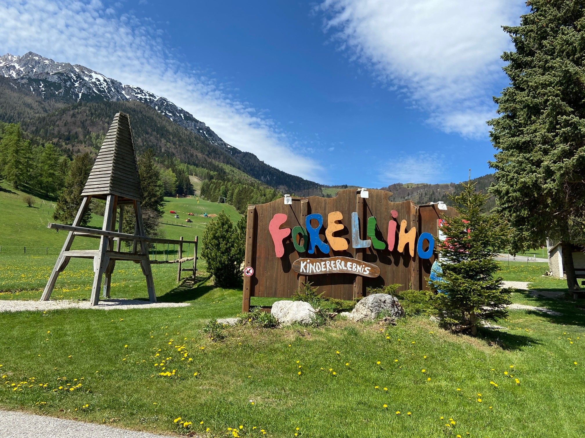 Eingang zum Forellino Kinderspielplatz mit Berglandschaft im Hintergrund.