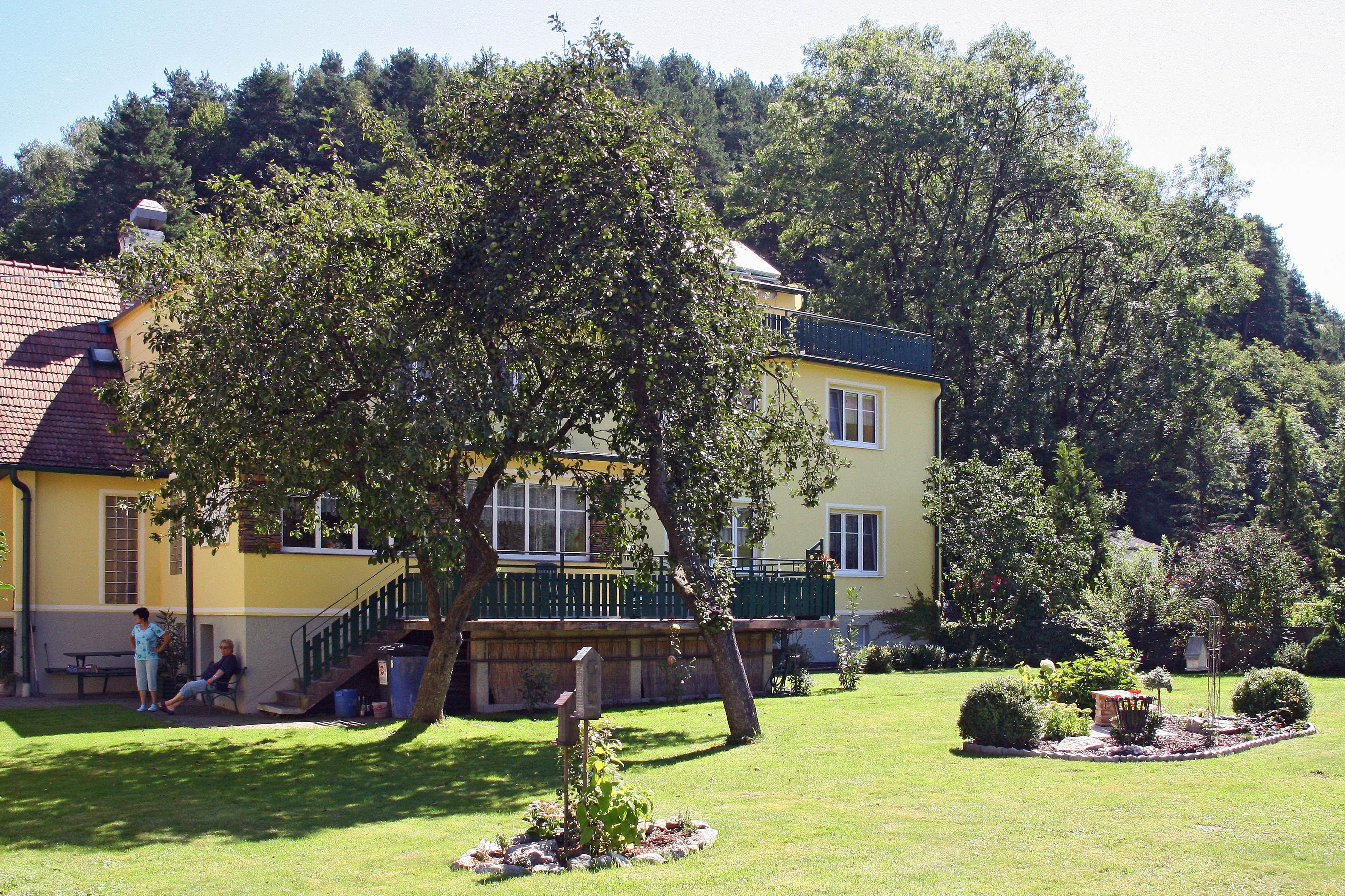 Gelbes Haus mit Garten und Bäumen, Personen sitzen im Schatten.
