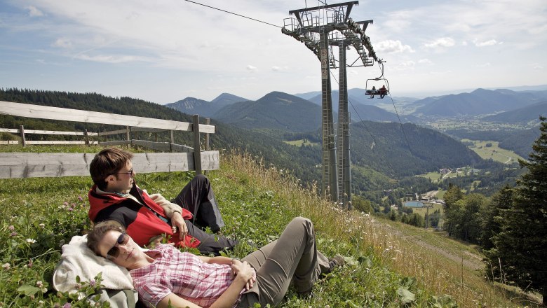 Zwei Personen entspannen auf einer Wiese neben einer Sesselbahn mit Bergblick.