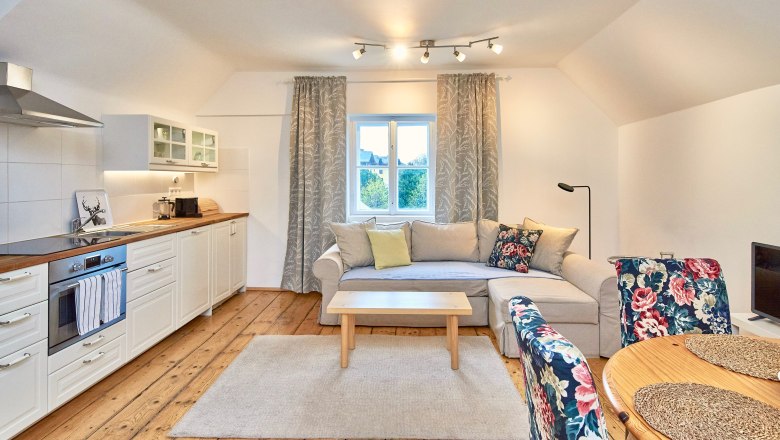 Gemütliches Wohnzimmer mit offener Küche, Holzboden, beiger Couch, floral gemusterten Stühlen und einem Fenster mit Vorhängen.