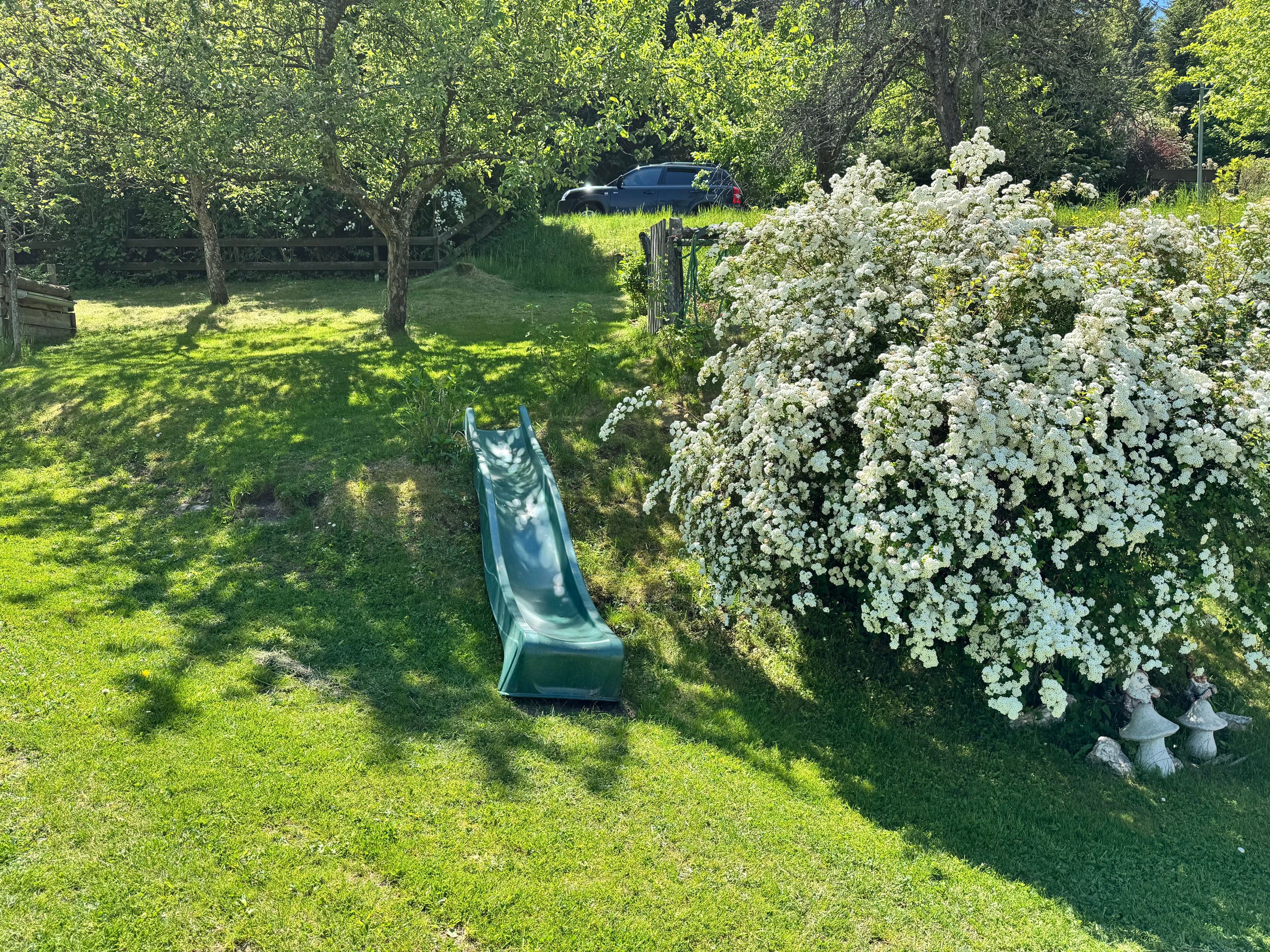 Grüne Rutsche auf einem Hügel im Garten, umgeben von Bäumen und blühenden Sträuchern.