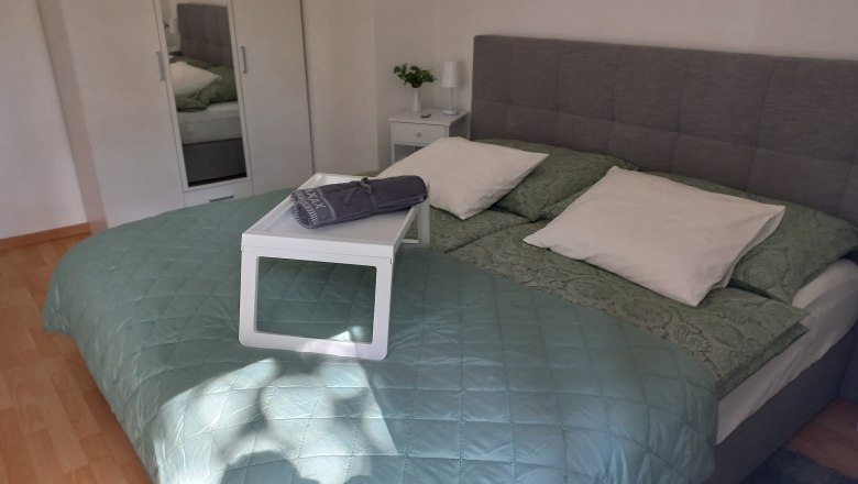 Ein gem&uuml;tliches Schlafzimmer mit einem gro&szlig;en Bett, gr&uuml;ner Bettdecke und wei&szlig;en Kissen. Ein Tablett mit Handtuch liegt auf dem Bett.
