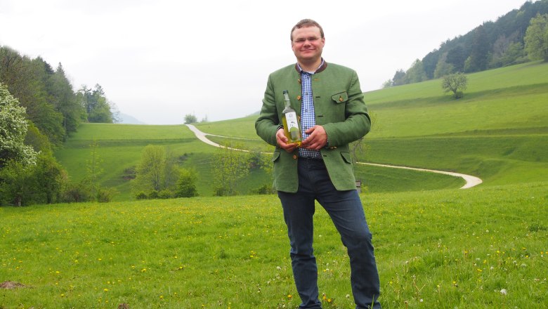 Mann in grüner Jacke hält eine Flasche auf einer grünen Wiese mit Hügeln im Hintergrund.
