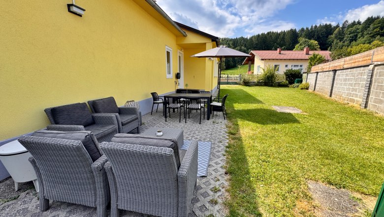 Terrasse mit Gartenmöbeln und Sonnenschirm neben gelbem Haus.