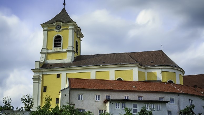 Gelbe Kirche mit Turm und roten Dächern vor bewölktem Himmel.