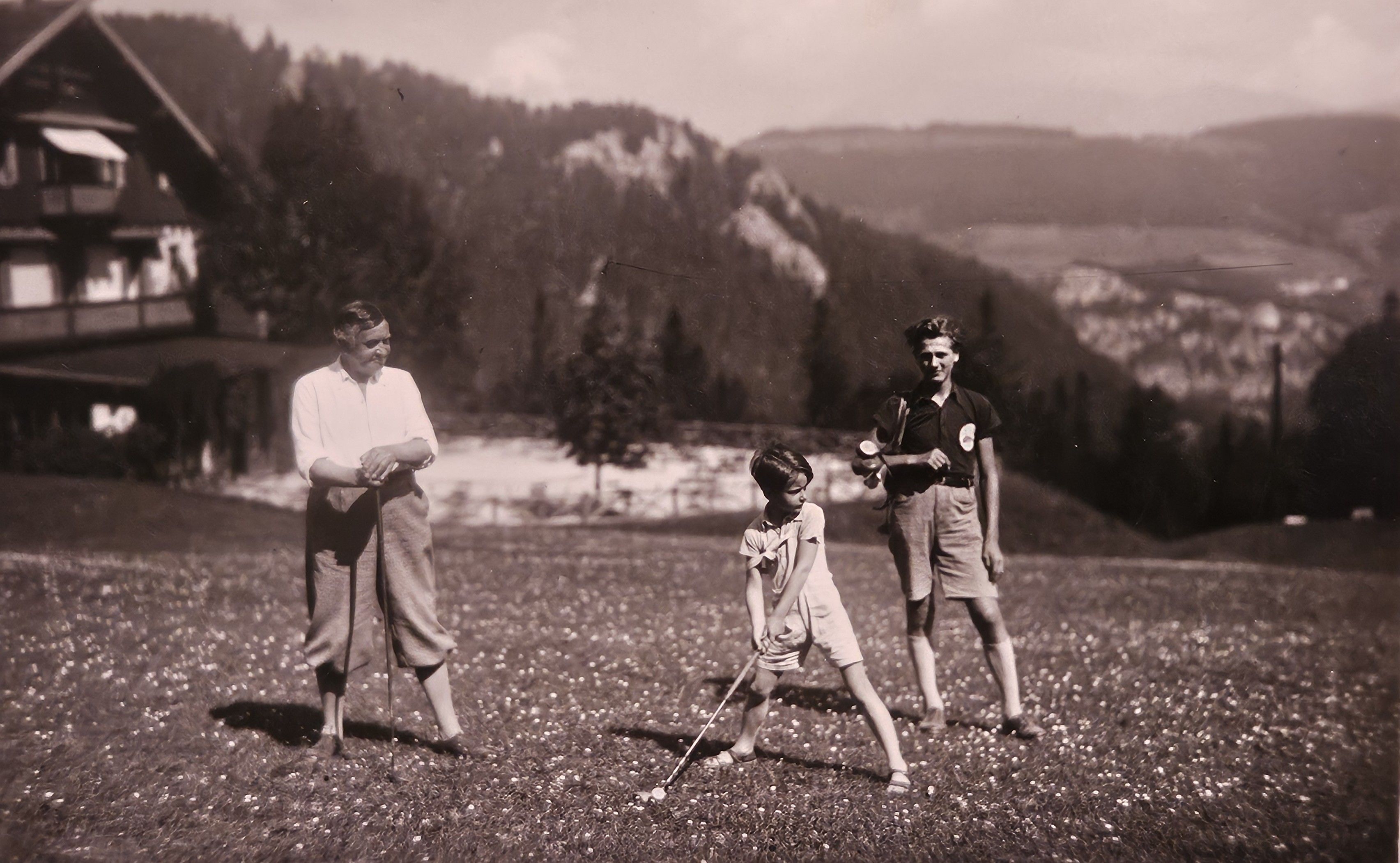 Ein Erwachsener und zwei Kinder spielen auf einem Golfplatz in alpiner Landschaft und einem Gebäude im Hintergrund.