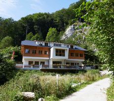 Hütte liegt zwischen vielen Bäumen am Beginn einer Felsschlucht, im Vordergrund ist ein Weg.