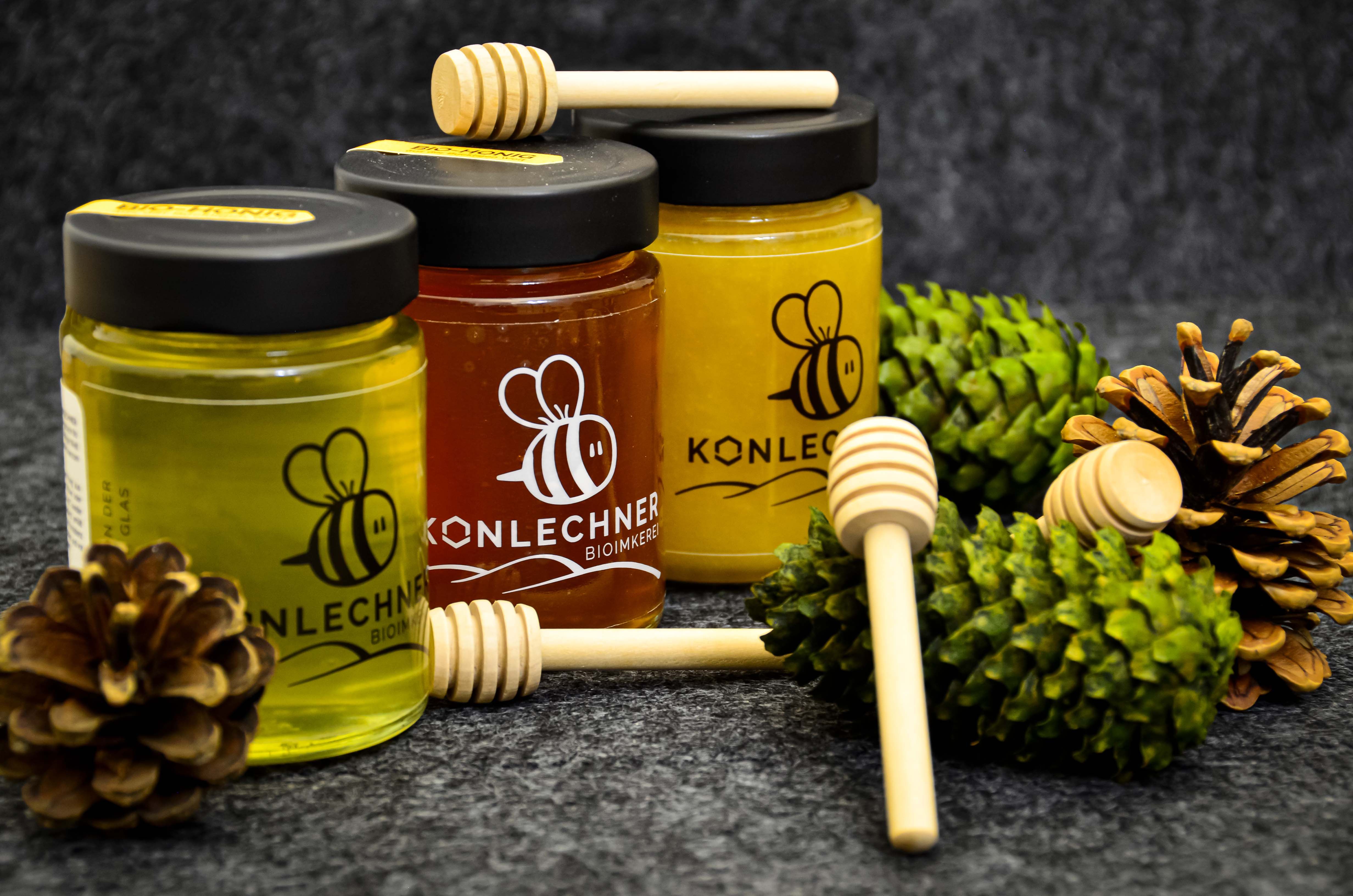 Drei verschiedene Honigsorten in Gläsern vor dunkelgrauem Hintergrund, umgeben von Tannenzapfen und Honiglöffeln.