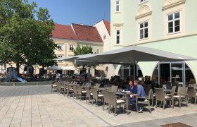 Ein Schanigarten vor dem Eterno mit Sonnenschirmen und Sitzpl&auml;tzen auf einem gepflasterten Platz.