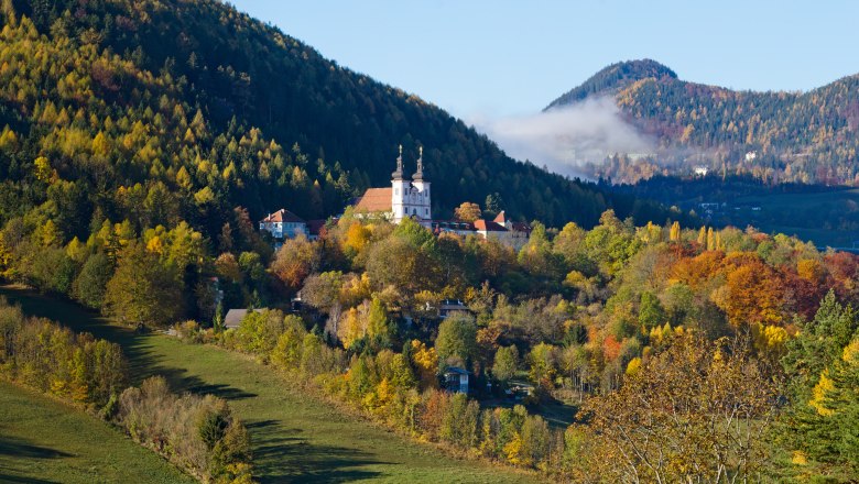 Herbstliche Landschaft mit Kirche in Maria Schutz, umgeben von bunten Bäumen und Hügeln.