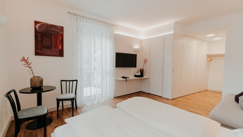 Modern, bright hotel room with double bed, TV, table and chairs, wardrobe and decorative picture on the wall.