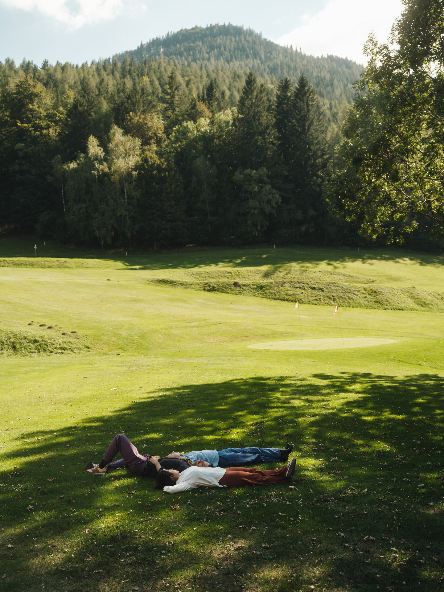 Unter dem sanften Licht der Nachmittagssonne entspannen drei Freunde auf der grünen Wiese, umgeben von der majestätischen Kulisse der Wiener Alpen. Die ruhige Atmosphäre und das sanfte Rascheln der Blätter laden dazu ein, die Seele baumeln zu lassen und die Schönheit der Natur zu genießen.