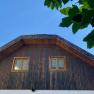 Attic of a house with wooden paneling and two windows, surrounded by green leaves and blue sky.