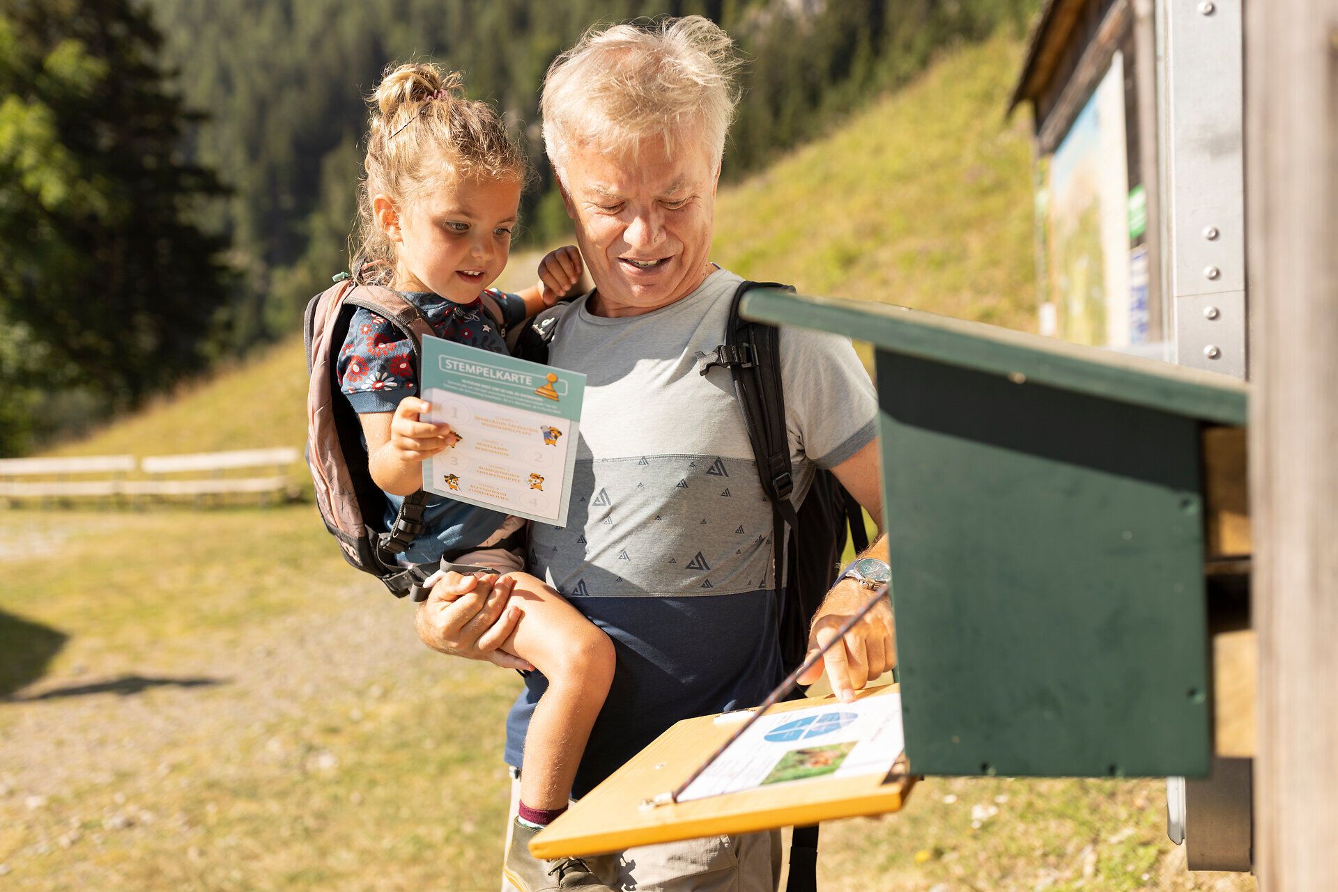 Ein fröhlicher Großvater hält seine Enkelin in den Armen, während sie gemeinsam an einer Informationsstelle stehen. Die malerische Umgebung der Wiener Alpen lädt zu unvergesslichen Wanderabenteuern ein, die Familien zusammenbringen und die Schönheit der Natur erlebbar machen.